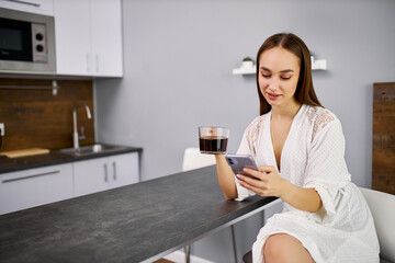 smiling caucasian lady looks at screen of smartphone and drinks coffee, young woman spend her morning alone, holding mobile phone