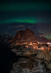 Fototapeta na wymiar A vertical shot of aurora borealis over illuminated fishing village with rorbu houses in Homnoya, Lofoten island, Norway