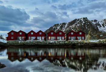 Obraz premium A horizontal shot of a symmetric row of classic red rorbu next to snowy mountain ranges reflecting water in Ballstad, Lofoten island, Norway