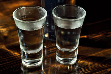 Frozen glasses with alcohol near the bottle. Drinks on a dark brown table. Transparent utensils on a black background. Old shabby boards under glass.