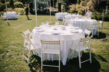 Banquet table on a green lawn. White wooden chairs. Rack and cutlery. Floral arrangement of pink flowers. On the table is a white tablecloth.