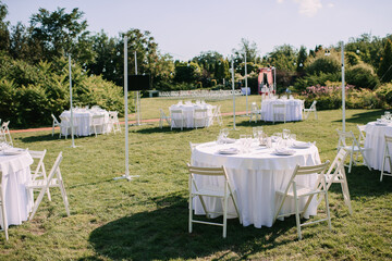 Banquet table on a green lawn. White wooden chairs. Rack and cutlery. Floral arrangement of pink flowers. On the table is a white tablecloth.