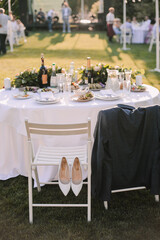 Banquet table on a green lawn. White wooden chairs. Rack and cutlery. Floral arrangement of pink flowers. On the table is a white tablecloth.