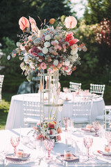 Banquet table on a green lawn. Rack and cutlery. Floral arrangement of pink flowers. Transparent banquet chairs. On the table is a white tablecloth.