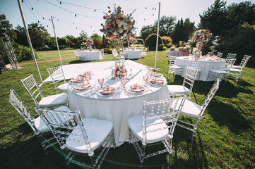 Banquet table on a green lawn. Rack and cutlery. Floral arrangement of pink flowers. Transparent banquet chairs. On the table is a white tablecloth.