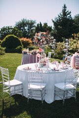 Banquet table on a green lawn. Rack and cutlery. Floral arrangement of pink flowers. Transparent banquet chairs. On the table is a white tablecloth.