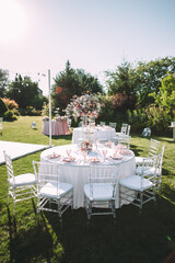 Fototapeta premium Banquet table on a green lawn. Rack and cutlery. Floral arrangement of pink flowers. Transparent banquet chairs. On the table is a white tablecloth.