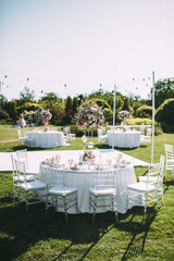 Banquet table on a green lawn. Rack and cutlery. Floral arrangement of pink flowers. Transparent banquet chairs. On the table is a white tablecloth.