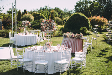 Banquet table on a green lawn. Rack and cutlery. Floral arrangement of pink flowers. Transparent banquet chairs. On the table is a white tablecloth.