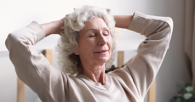 Head shot peaceful middle aged retired hoary woman folded hands behind head, recollecting positive life moments. Close up happy beautiful granny enjoying free lazy time, relaxing alone at home.