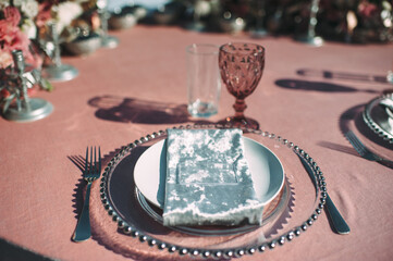 Banquet table on a green lawn. Racks and cutlery, velvet napkins, pink glasses. Floral arrangement of pink flowers. Silver candlesticks and candles. On the table is a white tablecloth.