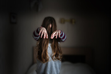 A little girl with long hair is standing in the room like a zombie. A child with outstretched arms wanders around the room. A terrible frightening children's concept.