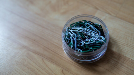 Top view of full paper clips in a transparent box on wooden background. It is device used to hold sheets of paper together made of steel wire bent to a looped shape. Selective focus.