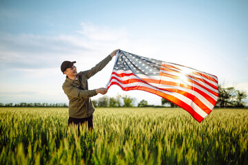 Young man wearing green shirt and cap lets the american flag fly on the wind at the green wheat field. Patriotic boy celebrates usa independence day on 4th of July with a national flag in his hands.