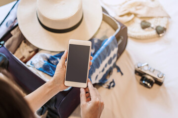 Young asian woman sitting on the bed using smartphone and packing her suitcase preparing for travel on summer vacation