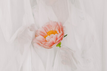 Aesthetic sensual image of beautiful woman behind tulle holding pink peony. Stylish girl gently holding peony flower in hand with jewelry. Soft image