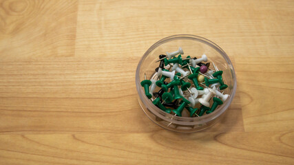 Top view of full thumb tacks in a transparent box on wooden background. It is a short nail or pin used to fasten items to a wall or board for display. Selective focus on foreground.