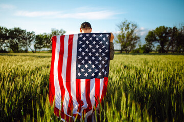 Young man wearing green shirt and cap stands wrapped in the american flag at the green wheat field. Patriotic boy celebrates usa independence day on the 4th of July with a national flag in his hands.