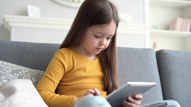 Curious cute preschool kid girl using digital tablet technology device sitting on sofa alone. Small child hold pad computer surfing internet play game at home. Children tech addiction concept