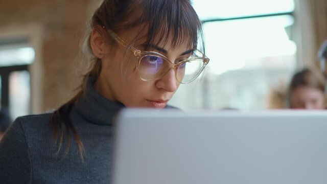 closeup face of Young business woman in eyeglasses concentrating on screen and typing on laptop while sitting at workplace or cafe. remote work, freelance, using laptop
