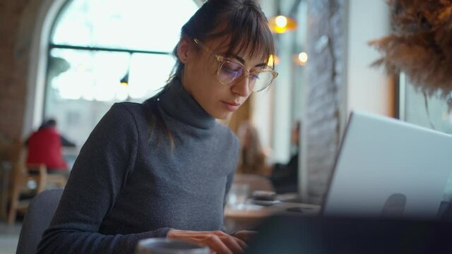 Portrait young woman in eyeglasses using laptop in coworking space or cafe and drincing coffee. female sitting in front open laptop computer. Study, learning, remote work, freelance.