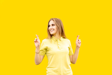 A young girl with fingers crossed hopes for good luck. Smiling with a white smile. Dressed in a yellow T-shirt. Isolated on yellow background.