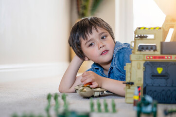 Portrait of School boy lying down on the carpet floor playing with soldiers, military car and figurine toys, Happy Kid playing wars and peace on his own at home, Children imagination and development