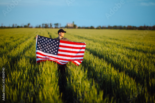 Young man wearing green shirt and cap lets the american flag fly on the wind at the green wheat field. Patriotic boy celebrates usa independence day on 4th of July with a national flag in his hands.