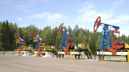 Fototapeta na wymiar Industrial oil well pump jacks pumping crude oil for fossil fuel energy with drilling rigs in oil field. Nodding donkey pumps working in middle of forest in sunny summer day. Oil mining machines.