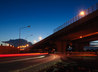 A view from underneath the highway with cars light trails passing by. The background has clear blue sky and big cloud.