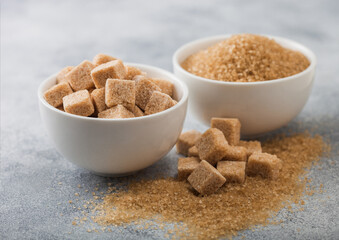 White bowl plates of natural brown sugar cubes and refined sugar on light table background.