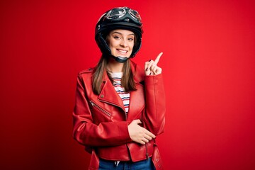 Young beautiful brunette motocyclist woman wearing motorcycle helmet and red jacket with a big smile on face, pointing with hand and finger to the side looking at the camera.
