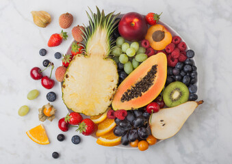 Fresh raw organic summer berries and exotic fruits in white plate on marble background. Pineapple, papaya, grapes, nectarine, orange, apricot, kiwi, pear, lychees, cherry and physalis.