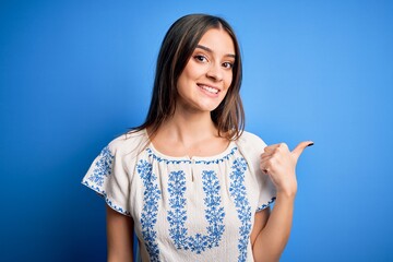 Young beautiful brunette woman wearing casual t-shirt standing over blue background smiling with happy face looking and pointing to the side with thumb up.