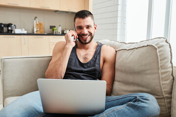 Joyful young man talking on cellphone and using laptop