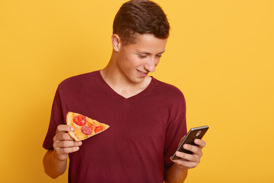 Photo of busy curious joyful teenager holding his smartphone, using his device, checking social networking sites, having piece of pizza in one hand, wearing red t shirt. Youth and free time concept.