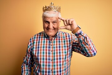 Senior handsome hoary man wearing golden crown of king over isolated yellow background smiling and confident gesturing with hand doing small size sign with fingers looking and the camera. Measure