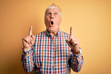 Naklejka premium Senior handsome hoary man wearing golden crown of king over isolated yellow background amazed and surprised looking up and pointing with fingers and raised arms.