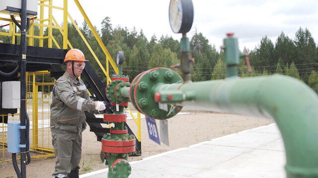 Oilman In Orange Helmet And Protective Glass Opening Or Closing Valve Of Wellhead. Injection Tree Is Destined For Injection Wells Sealing And Their Operating Regime Regulation. Oil Industry