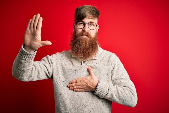 Handsome Irish redhead man with beard wearing casual sweater and glasses over red background Swearing with hand on chest and open palm, making a loyalty promise oath