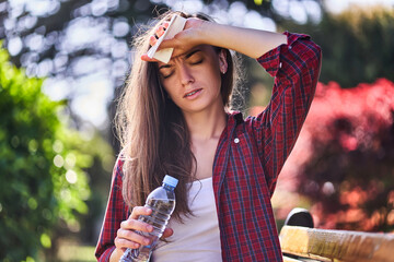 Tired sweating woman with refreshing water bottle hold on to forehead outdoors in hot summer weather
