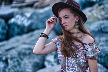 Portrait of attractive boho woman with blowing hair in the wind wearing a brown felt hat and gypsy bijouterie on the stones outdoors