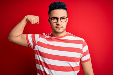 Young handsome man wearing casual striped t-shirt and glasses over isolated red background Strong person showing arm muscle, confident and proud of power