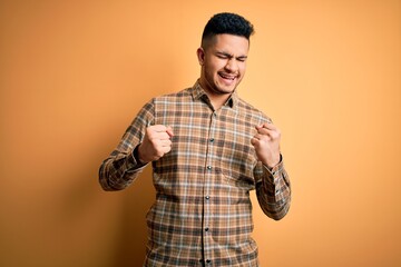 Young handsome man wearing casual shirt standing over isolated yellow background very happy and excited doing winner gesture with arms raised, smiling and screaming for success. Celebration concept.