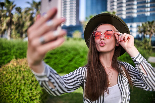 Gorgeous stylish casual modern carefree young hipster woman blogger wearing felt hat and bright red glasses blowing air kiss and using phone for taking a selfie photo portrait