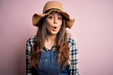 Young beautiful brunette farmer woman wearing apron and hat over pink background afraid and shocked with surprise expression, fear and excited face.