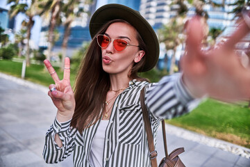 Gorgeous stylish casual modern carefree young hipster woman wearing felt hat and bright red glasses blowing air kiss and showing peace sign during taking selfie photo portrait