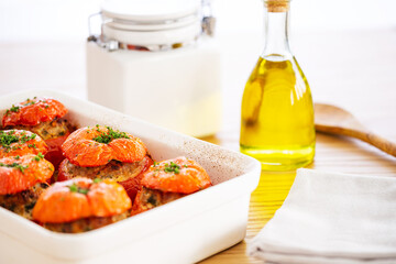 Mediterranean stuffed tomatoes with meat, bread crumbs, and herbs in a white oven dish, aside a bottle of olive oil, a white jar and a serving spoon, all on an oak wood table.