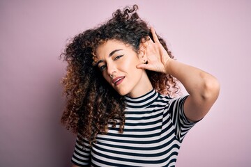 Young beautiful woman with curly hair and piercing wearing casual striped t-shirt smiling with hand over ear listening an hearing to rumor or gossip. Deafness concept.