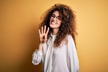 Young beautiful brunette woman with curly hair and piercing wearing shirt and glasses showing and pointing up with fingers number four while smiling confident and happy.